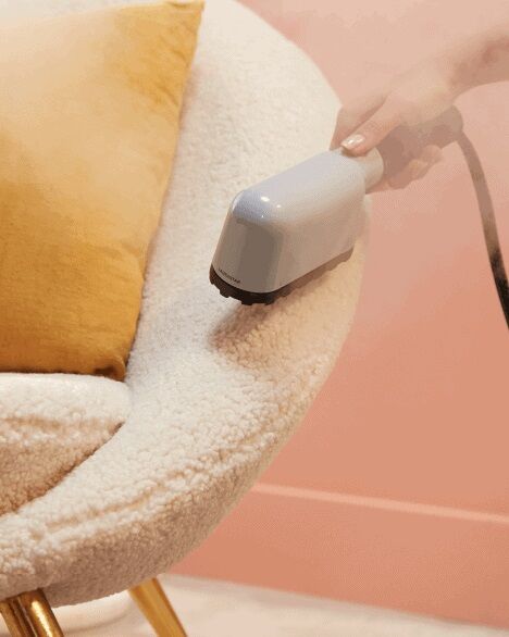 Person using a handheld steam cleaner on a plush white chair with a mustard yellow pillow