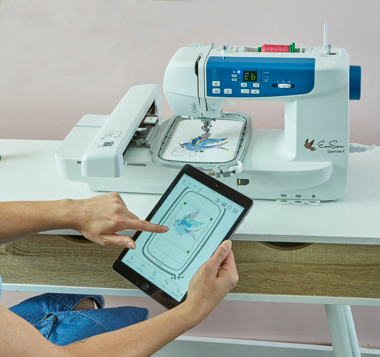 Person using a tablet to control an embroidery machine stitching a colorful bird design on fabric.