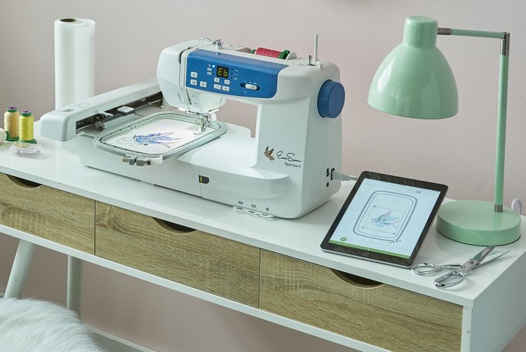 Embroidery machine working on a fabric with a tablet displaying the design, placed on a white desk with sewing tools