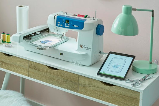 Embroidery machine working on a fabric with a tablet displaying the design, placed on a white desk with sewing tools