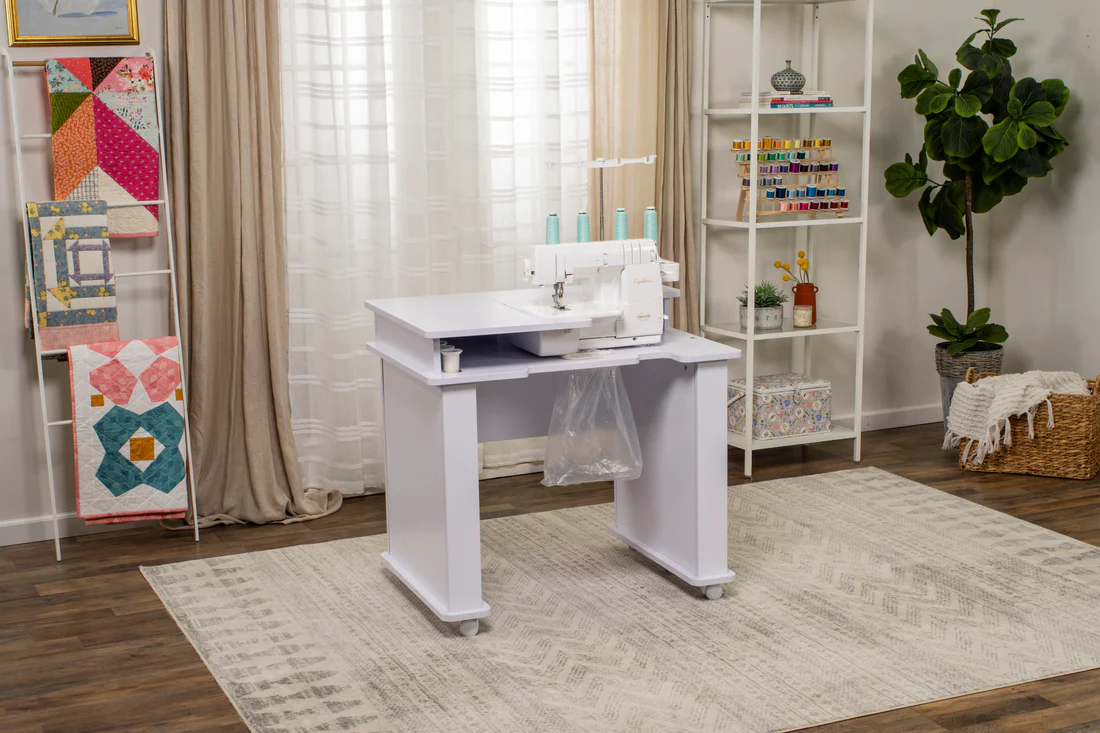 White serger sewing machine on a workstation table in a cozy sewing room with quilts and shelves