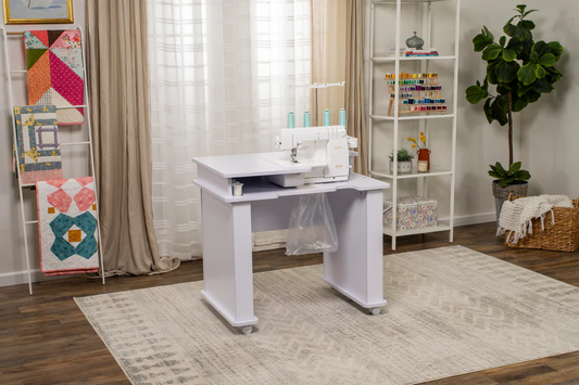 White serger sewing machine on a workstation table in a cozy sewing room with quilts and shelves