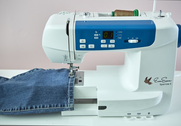 Close-up of a white and blue sewing machine stitching a pair of denim jeans, with brown thread loaded on top.