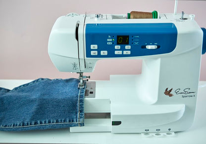 Close-up of a white and blue sewing machine stitching a pair of denim jeans, with brown thread loaded on top.