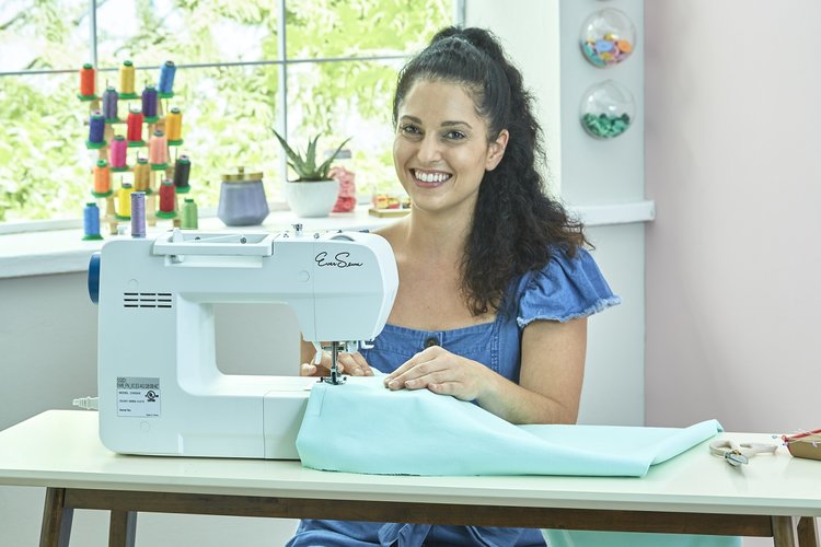 Smiling woman sewing fabric on a white sewing machine in a bright, colorful workspace with spools of thread and plants in the background