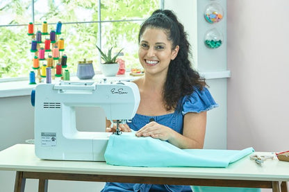 Smiling woman sewing fabric on a white sewing machine in a bright, colorful workspace with spools of thread and plants in the background